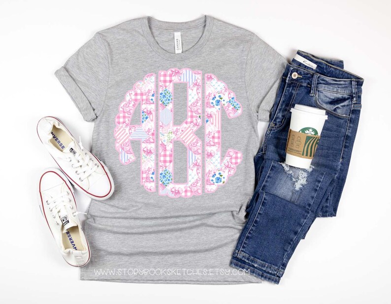 May include: A gray t-shirt with a pink and blue patchwork monogram design. The monogram is in the center of the shirt. The shirt is laying on a white surface with a pair of white sneakers and a pair of blue jeans.