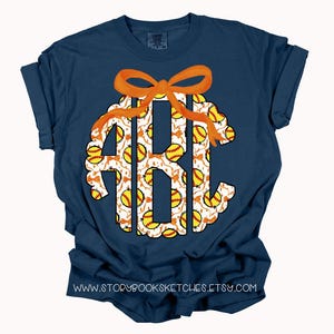 K&ouml;nnte beinhalten: Ein marineblaues T-Shirt mit einem Softball-Monogramm. Das Monogramm ist wei&szlig; mit einer orangefarbenen Schleife und einem Muster aus gelben und wei&szlig;en Softb&auml;llen.