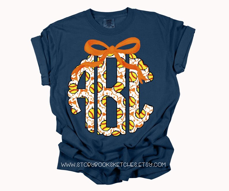 K&ouml;nnte beinhalten: Ein marineblaues T-Shirt mit einem Softball-Monogramm. Das Monogramm ist wei&szlig; mit einer orangefarbenen Schleife und einem Muster aus gelben und wei&szlig;en Softb&auml;llen.