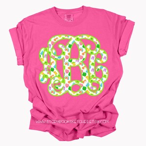May include: A bright pink t-shirt featuring a large monogram design. The monogram is outlined in green and filled with a pattern of rainbows, hearts, shamrocks, and other colorful shapes. The text "www.storybooksketches.etsy.com" is printed at the bottom.