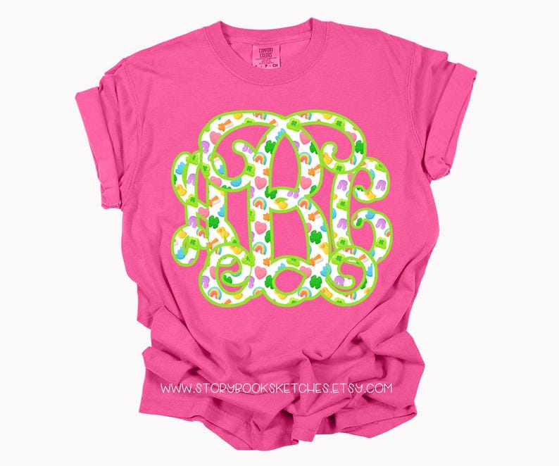 May include: A bright pink t-shirt featuring a large monogram design. The monogram is outlined in green and filled with a pattern of rainbows, hearts, shamrocks, and other colorful shapes. The text "www.storybooksketches.etsy.com" is printed at the bottom.