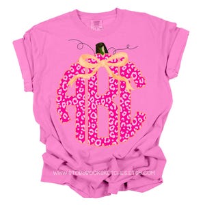 K&ouml;nnte beinhalten: Pinkes T-Shirt mit einem pinkfarbenen und wei&szlig;en Leopardenmuster-K&uuml;rbis, der mit den Buchstaben "ABC" monogrammiert ist. Der K&uuml;rbis hat eine goldene Schleife.