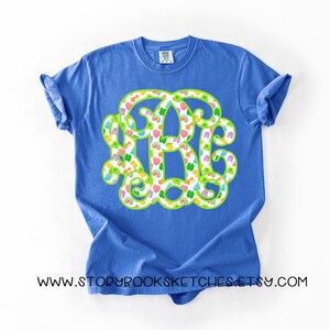 May include: A blue t-shirt featuring a large monogram design. The monogram is outlined in green and filled with a pattern of colorful hearts, rainbows, and clovers. The text "WWW.STORYBOOKSKETCHES.ETSY.COM" is printed at the bottom of the shirt.