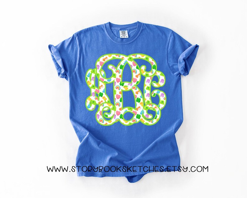 May include: A blue t-shirt featuring a large monogram design. The monogram is outlined in green and filled with a pattern of colorful hearts, rainbows, and clovers. The text "WWW.STORYBOOKSKETCHES.ETSY.COM" is printed at the bottom of the shirt.