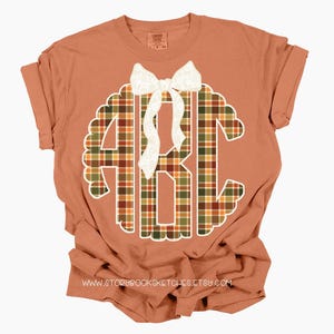 Puede incluir: Una camiseta de color terracota con un gran dise&ntilde;o de monograma. El monograma es un patr&oacute;n de cuadros en tonos verdes, naranjas y marrones, con un borde festoneado blanco y un lazo blanco. El texto "www.storybooksketches.etsy.com" est&aacute; en la parte inferior.