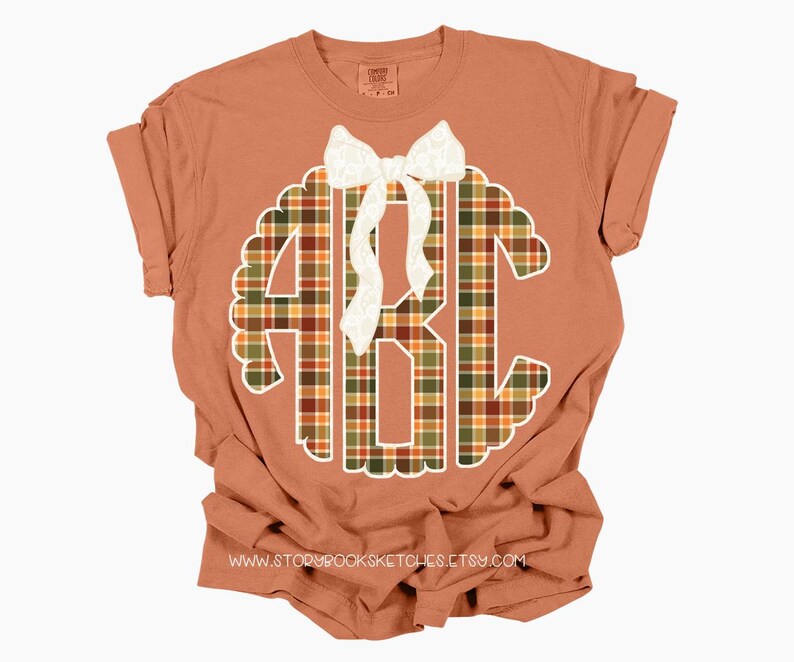 Puede incluir: Una camiseta de color terracota con un gran dise&ntilde;o de monograma. El monograma es un patr&oacute;n de cuadros en tonos verdes, naranjas y marrones, con un borde festoneado blanco y un lazo blanco. El texto "www.storybooksketches.etsy.com" est&aacute; en la parte inferior.