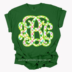May include: Green t-shirt with a large, monogram-style design in white, outlined in green. The design is filled with colorful shamrocks, rainbows, and hearts. The text "www.storybooksketches.etsy.com" is printed at the bottom.