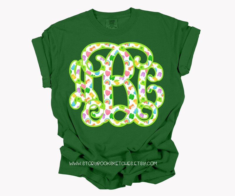May include: Green t-shirt with a large, monogram-style design in white, outlined in green. The design is filled with colorful shamrocks, rainbows, and hearts. The text "www.storybooksketches.etsy.com" is printed at the bottom.