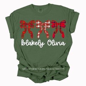 May include: A green t-shirt with a red and white plaid bow design and the names "Blakely Olivia" printed in white.