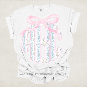 Puede incluir: Camiseta blanca con un lazo rosa y un monograma de las letras A, B y C. El monograma est&aacute; rodeado de un patr&oacute;n floral en azul y rosa.