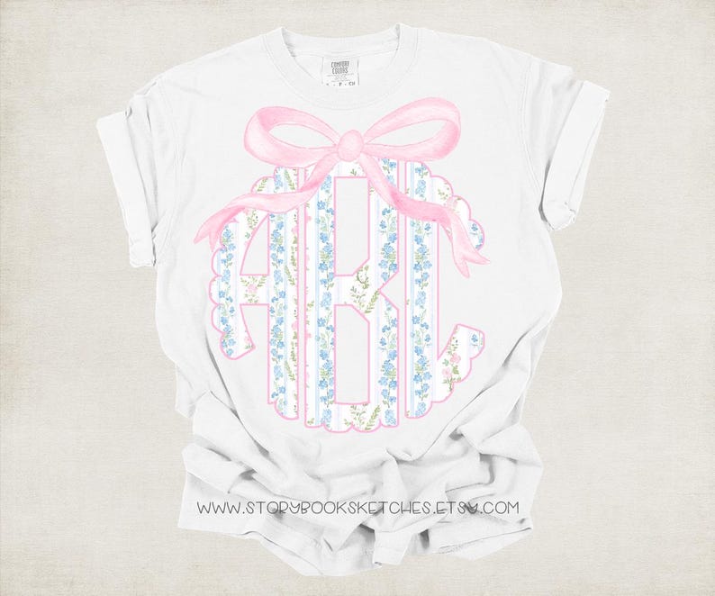 Puede incluir: Camiseta blanca con un lazo rosa y un monograma de las letras A, B y C. El monograma est&aacute; rodeado de un patr&oacute;n floral en azul y rosa.