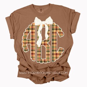 Puede incluir: Una camiseta marr&oacute;n con un monograma blanco festoneado con las letras ABC en un patr&oacute;n de cuadros naranja, verde y marr&oacute;n. Un lazo blanco est&aacute; colocado en la parte superior del monograma. La direcci&oacute;n del sitio web www.storybooksketches.com est&aacute; impresa en la parte inferior.
