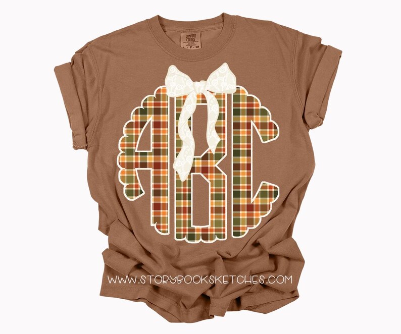 Puede incluir: Una camiseta marr&oacute;n con un monograma blanco festoneado con las letras ABC en un patr&oacute;n de cuadros naranja, verde y marr&oacute;n. Un lazo blanco est&aacute; colocado en la parte superior del monograma. La direcci&oacute;n del sitio web www.storybooksketches.com est&aacute; impresa en la parte inferior.