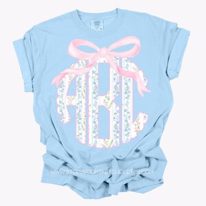 Puede incluir: Camiseta azul claro con un lazo rosa y un monograma en un patr&oacute;n floral. El monograma est&aacute; en un c&iacute;rculo blanco con un patr&oacute;n floral azul y blanco.