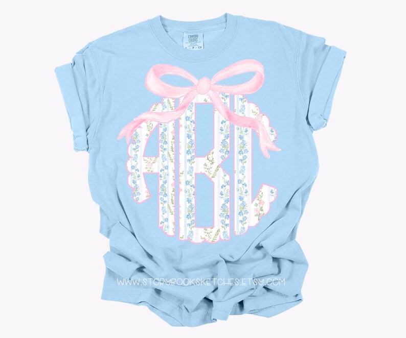Puede incluir: Camiseta azul claro con un lazo rosa y un monograma en un patr&oacute;n floral. El monograma est&aacute; en un c&iacute;rculo blanco con un patr&oacute;n floral azul y blanco.