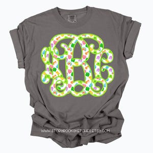 May include: A gray t-shirt featuring a large, green-outlined monogram with a pattern of hearts, rainbows, and shamrocks. The shirt has rolled-up sleeves and the website address www.storybooksketches.etsy.com is printed at the bottom.