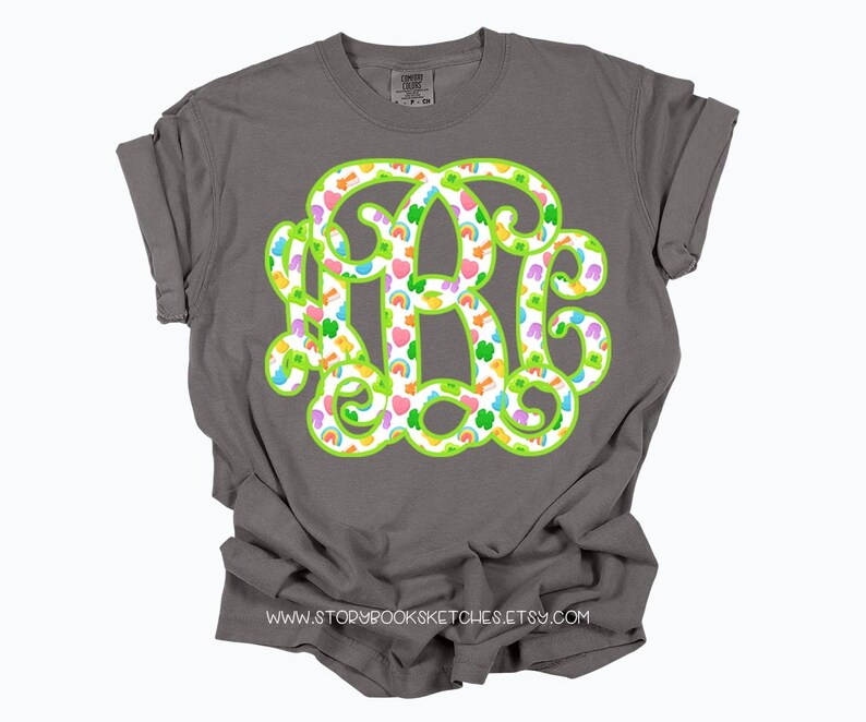 May include: A gray t-shirt featuring a large, green-outlined monogram with a pattern of hearts, rainbows, and shamrocks. The shirt has rolled-up sleeves and the website address www.storybooksketches.etsy.com is printed at the bottom.