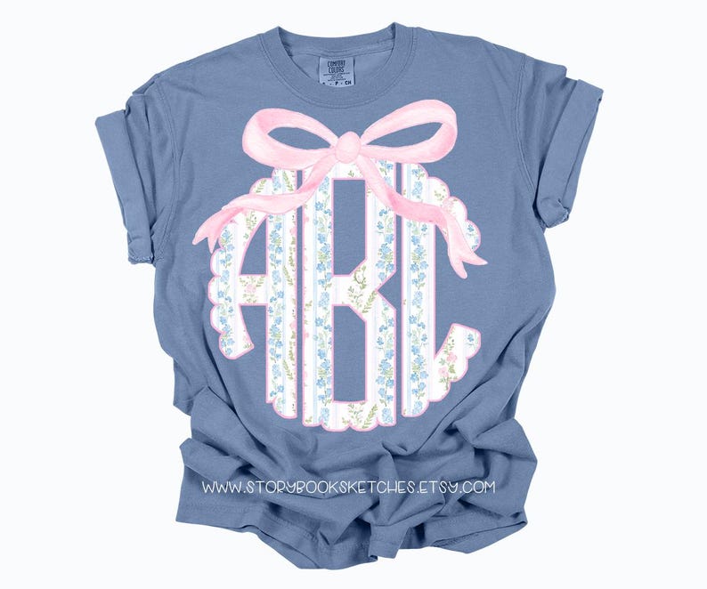 Puede incluir: Una camiseta azul con un lazo rosa y un monograma en un patr&oacute;n floral. El monograma est&aacute; en un c&iacute;rculo con un borde festoneado.