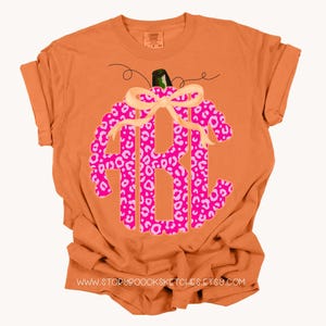 K&ouml;nnte beinhalten: Orangefarbenes T-Shirt mit einem rosa-wei&szlig;en Leopardenmuster-K&uuml;rbis mit Schleife. Der K&uuml;rbis hat ein Monogramm in der Mitte. Der Text "www.storybooksketches.etsy.com" ist auf der Unterseite des Shirts aufgedruckt.