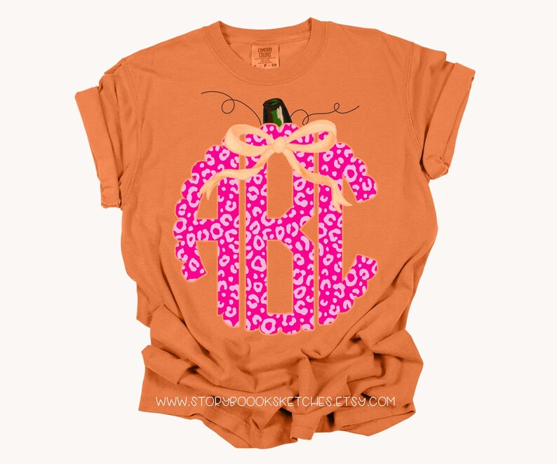 K&ouml;nnte beinhalten: Orangefarbenes T-Shirt mit einem rosa-wei&szlig;en Leopardenmuster-K&uuml;rbis mit Schleife. Der K&uuml;rbis hat ein Monogramm in der Mitte. Der Text "www.storybooksketches.etsy.com" ist auf der Unterseite des Shirts aufgedruckt.