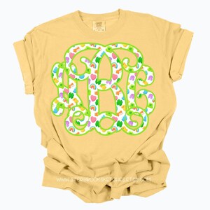 May include: A yellow t-shirt featuring a large monogram design. The monogram is outlined in green and filled with colorful shamrocks, rainbows, and hearts. The shirt has rolled-up sleeves and a crew neck.