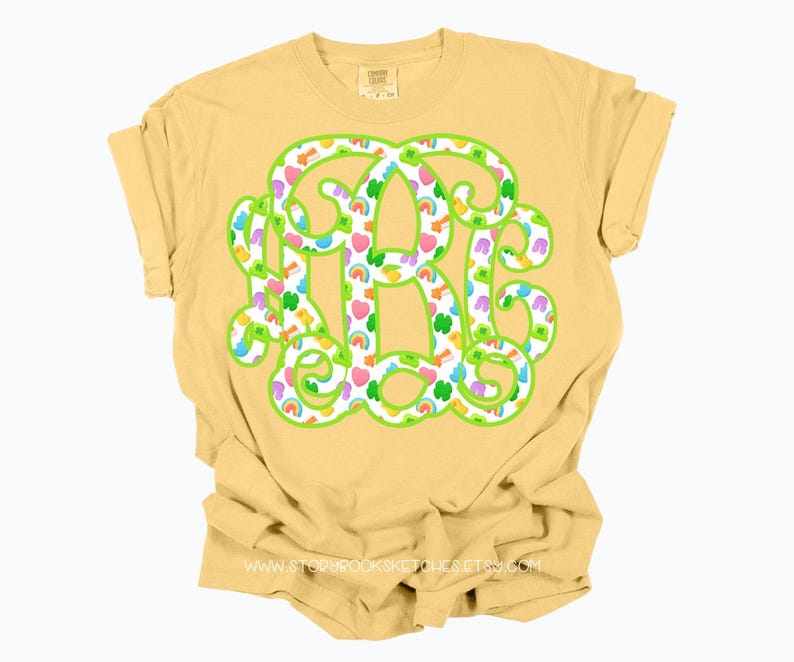 May include: A yellow t-shirt featuring a large monogram design. The monogram is outlined in green and filled with colorful shamrocks, rainbows, and hearts. The shirt has rolled-up sleeves and a crew neck.