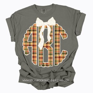 Puede incluir: Camiseta gris con mangas enrolladas con un monograma festoneado blanco con las letras ABC en un patr&oacute;n de cuadros. Un lazo blanco est&aacute; centrado encima del monograma. La direcci&oacute;n del sitio web www.storybooksketches.etsy.com est&aacute; impresa en la parte inferior.