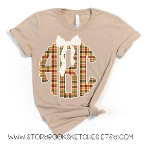 Puede incluir: Camiseta color beige con un dise&ntilde;o de monograma a cuadros en colores oto&ntilde;ales, con las letras ABC. Un lazo blanco est&aacute; colocado encima del monograma. La direcci&oacute;n del sitio web www.storybooksketches.etsy.com est&aacute; impresa en la parte inferior de la camiseta.