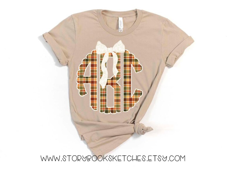 Puede incluir: Camiseta color beige con un dise&ntilde;o de monograma a cuadros en colores oto&ntilde;ales, con las letras ABC. Un lazo blanco est&aacute; colocado encima del monograma. La direcci&oacute;n del sitio web www.storybooksketches.etsy.com est&aacute; impresa en la parte inferior de la camiseta.