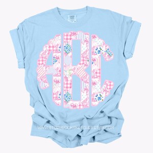 May include: A light blue t-shirt with a pink and blue patchwork monogram. The monogram is in the shape of a circle and features a variety of floral and geometric patterns.