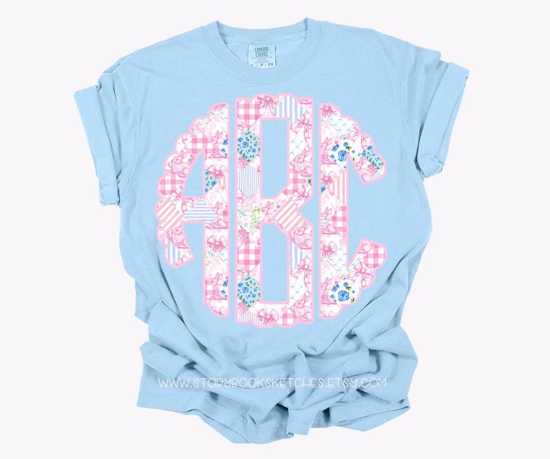 May include: A light blue t-shirt with a pink and blue patchwork monogram. The monogram is in the shape of a circle and features a variety of floral and geometric patterns.