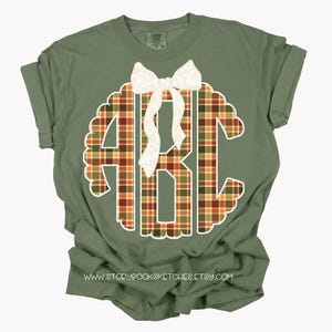 Puede incluir: Camiseta verde oliva con un gran monograma a cuadros en el centro. El monograma, con las letras "ABC", est&aacute; delineado en blanco. Un lazo blanco est&aacute; colocado en la parte superior del monograma. La direcci&oacute;n del sitio web www.storybooksketches.etsy.com est&aacute; impresa en la parte inferior.