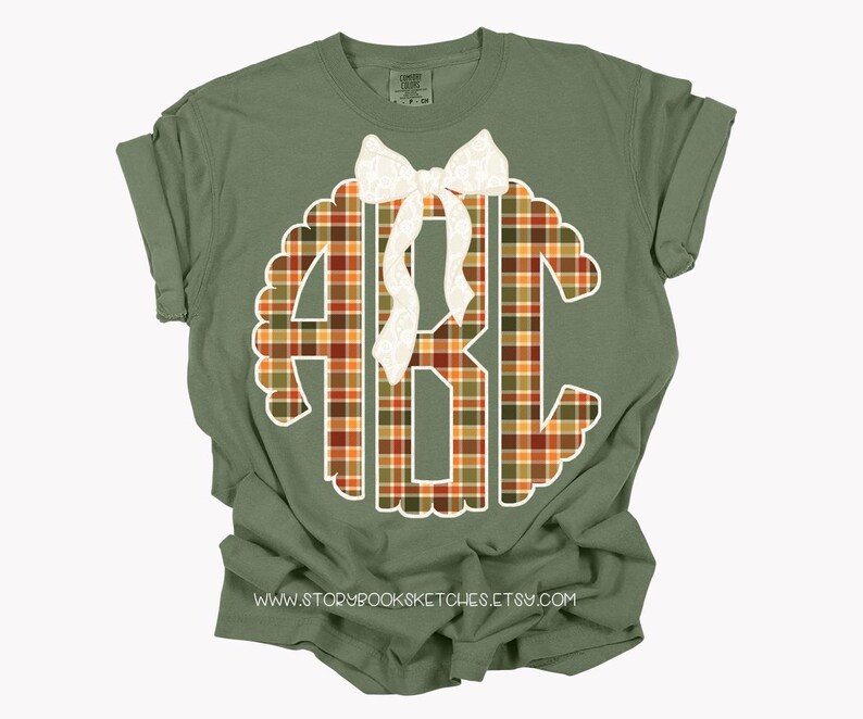 Puede incluir: Camiseta verde oliva con un gran monograma a cuadros en el centro. El monograma, con las letras "ABC", est&aacute; delineado en blanco. Un lazo blanco est&aacute; colocado en la parte superior del monograma. La direcci&oacute;n del sitio web www.storybooksketches.etsy.com est&aacute; impresa en la parte inferior.