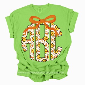 K&ouml;nnte beinhalten: Ein limegr&uuml;nes T-Shirt mit einem Softball-Monogramm-Design. Das Monogramm ist von einem wei&szlig;en und orangefarbenen Softball-Muster umgeben. Das Monogramm ist mit einer orangefarbenen Schleife verziert.