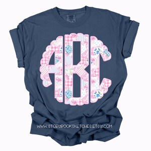 May include: A navy blue t-shirt with a pink and white floral monogram. The monogram is in the shape of a circle with a scalloped edge. The t-shirt has a crew neck and short sleeves.