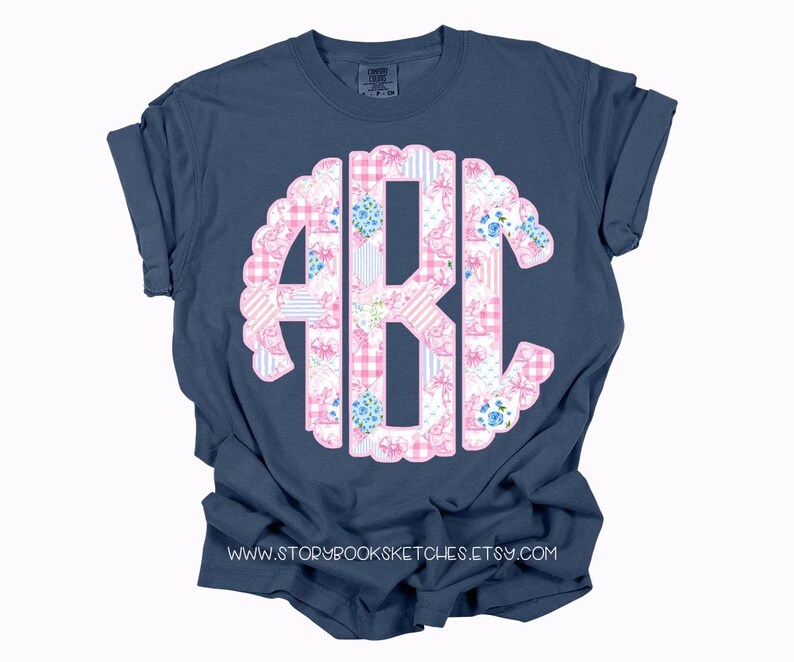 May include: A navy blue t-shirt with a pink and white floral monogram. The monogram is in the shape of a circle with a scalloped edge. The t-shirt has a crew neck and short sleeves.