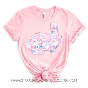 Pu&ograve; includere: Una maglietta rosa chiaro con un design a coniglio patchwork. Il coniglio &egrave; composto da vari motivi floreali e a quadri in rosa, blu e bianco. Il testo "WWW.STORYBOOKSKETCHES.ETSY.COM" &egrave; in basso.