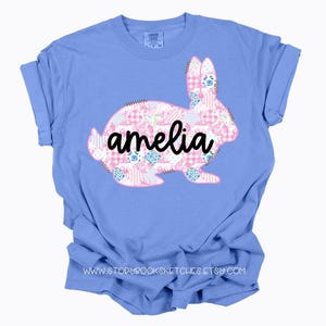 Pu&ograve; includere: Una maglietta azzurra con un design a coniglio patchwork in motivi floreali e gingham rosa, bianchi e blu. Il nome "amelia" &egrave; scritto in corsivo nero sul coniglio. La maglietta ha le maniche arrotolate.