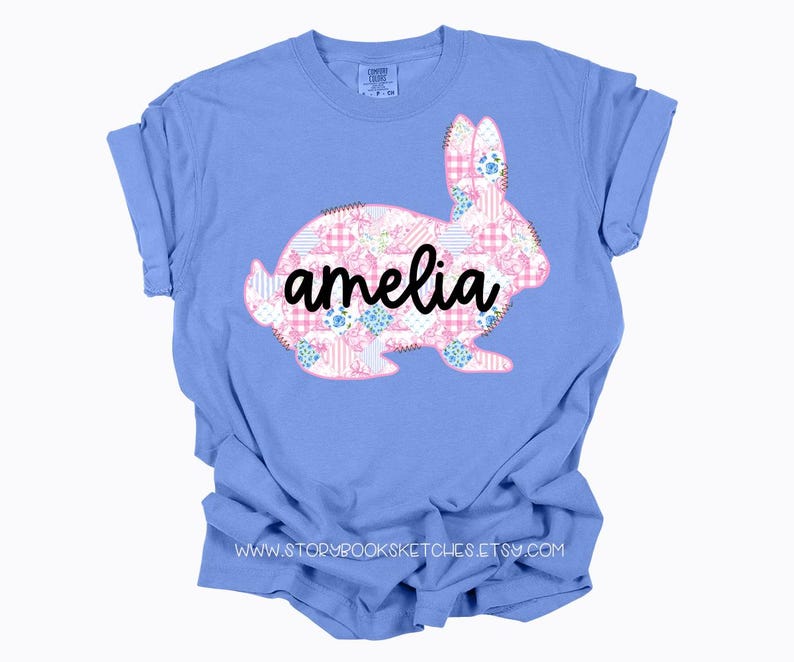 Pu&ograve; includere: Una maglietta azzurra con un design a coniglio patchwork in motivi floreali e gingham rosa, bianchi e blu. Il nome "amelia" &egrave; scritto in corsivo nero sul coniglio. La maglietta ha le maniche arrotolate.