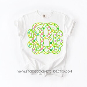 May include: White t-shirt featuring a large, green monogram design filled with colorful hearts, rainbows, and shamrocks. The shirt has a classic crew neck and short sleeves. The text "WWW.STORYBOOKSKETCHES.ETSY.COM" is printed at the bottom of the shirt.