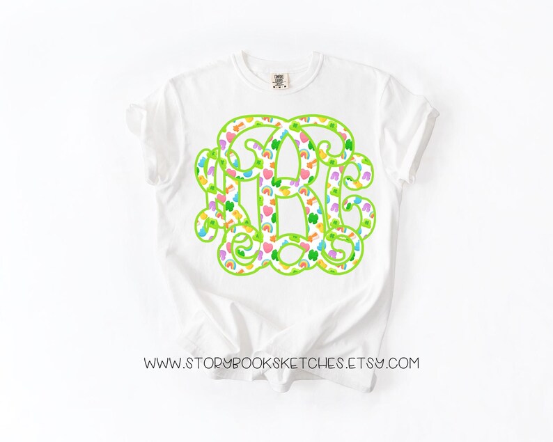 May include: White t-shirt featuring a large, green monogram design filled with colorful hearts, rainbows, and shamrocks. The shirt has a classic crew neck and short sleeves. The text "WWW.STORYBOOKSKETCHES.ETSY.COM" is printed at the bottom of the shirt.