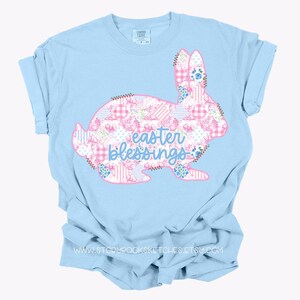 Pu&ograve; includere: T-shirt azzurra con un disegno di coniglio con un motivo patchwork di fiori rosa e bianchi e quadretti. Le parole "Easter Blessings" sono in blu. Una maglietta dallo stile casual e confortevole.