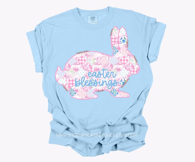 Pu&ograve; includere: T-shirt azzurra con un disegno di coniglio con un motivo patchwork di fiori rosa e bianchi e quadretti. Le parole "Easter Blessings" sono in blu. Una maglietta dallo stile casual e confortevole.
