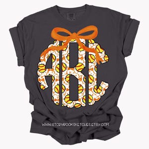 K&ouml;nnte beinhalten: Ein dunkelgraues T-Shirt mit einem wei&szlig;en Monogramm, das mit einem Softball-Muster verziert ist. Das Monogramm ist mit einer orangefarbenen Schleife verziert. Der Text "www.storybooksketches.etsy.com" ist auf der Unterseite des T-Shirts aufgedruckt.