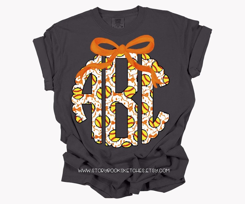 K&ouml;nnte beinhalten: Ein dunkelgraues T-Shirt mit einem wei&szlig;en Monogramm, das mit einem Softball-Muster verziert ist. Das Monogramm ist mit einer orangefarbenen Schleife verziert. Der Text "www.storybooksketches.etsy.com" ist auf der Unterseite des T-Shirts aufgedruckt.