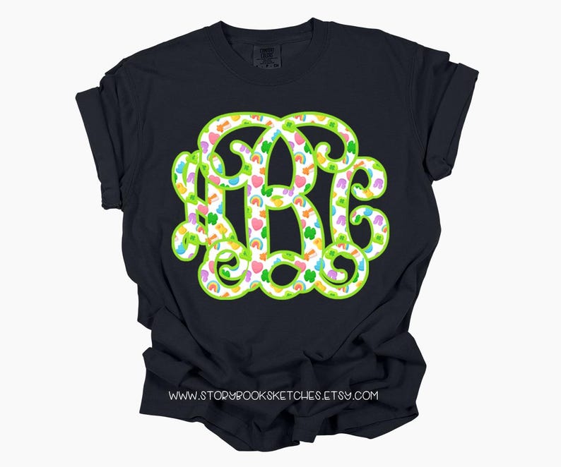 May include: A navy blue t-shirt featuring a large monogram design in the letters R, B, and C. The monogram is outlined in bright green and filled with a pattern of rainbows, hearts, and shamrocks. The website address www.storybooksketches.etsy.com is printed at the bottom.
