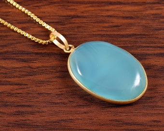 May include: A gold-coloured necklace featuring a light blue oval pendant. The pendant has a smooth, polished surface and is set in a gold-coloured bezel. The chain is a delicate, gold-coloured link chain.