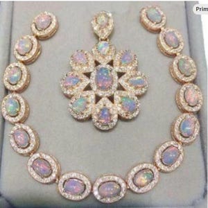 May include: A jewelry set featuring a necklace, pendant, and earrings. The set includes oval-shaped opals surrounded by small, clear gemstones, set in a gold-colored metal. The pendant is a flower shape, and the earrings are circular.