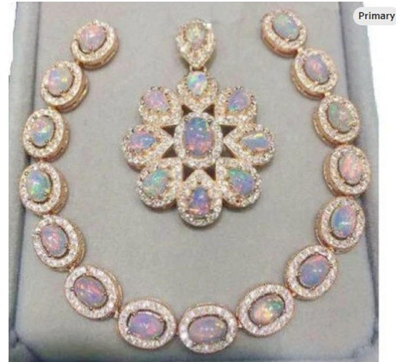 May include: A jewelry set featuring a necklace, pendant, and earrings. The set includes oval-shaped opals surrounded by small, clear gemstones, set in a gold-colored metal. The pendant is a flower shape, and the earrings are circular.