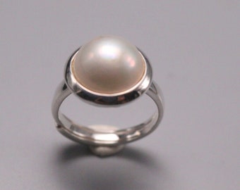 May include: A silver ring featuring a large, lustrous, off-white pearl. The pearl is set in a circular bezel, and the band is a simple, polished silver. The ring is displayed against a plain, light gray background.